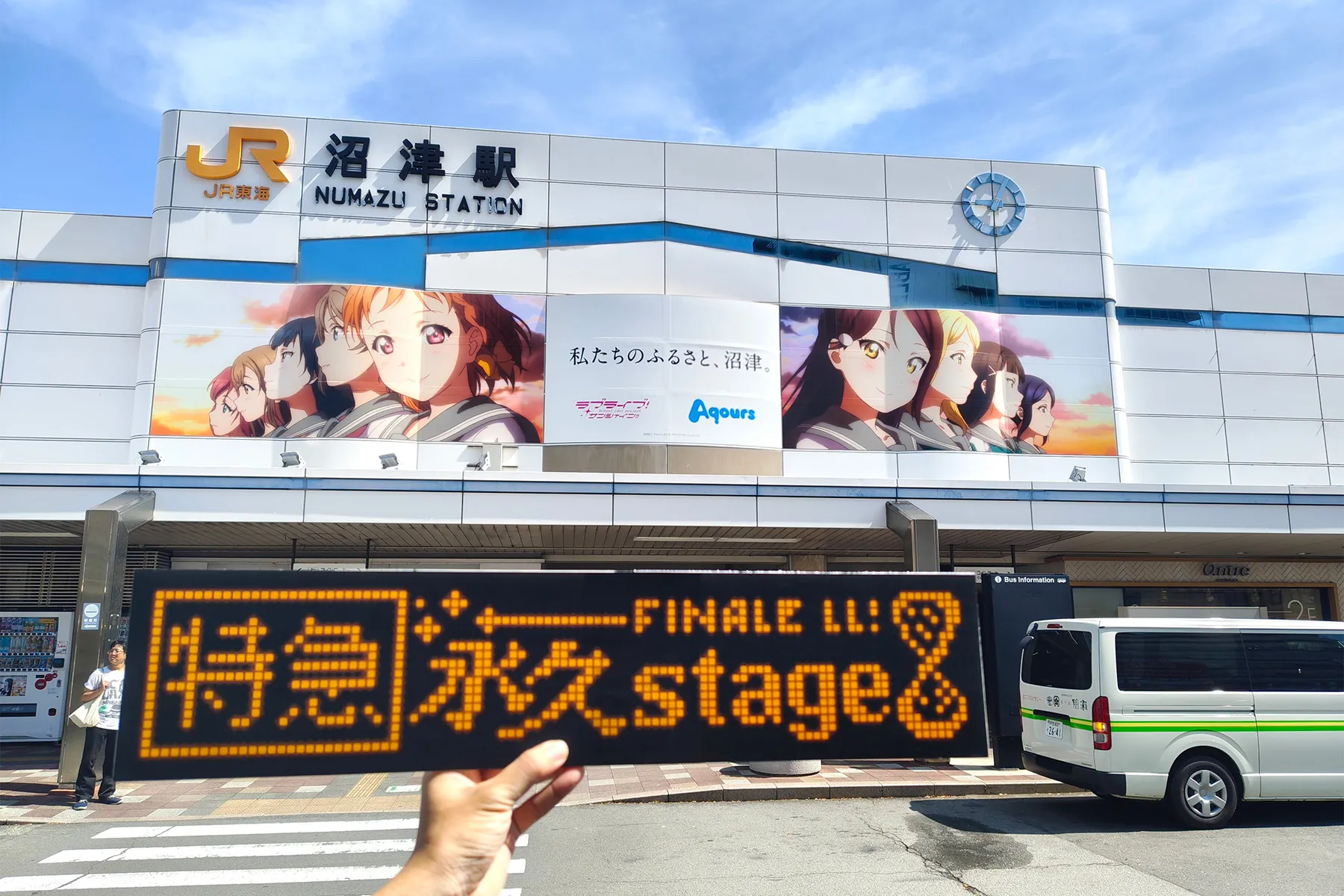 「【特急】永久stage」という文字が行先表示器風に描かれているボードと背景に沼津駅の南口駅舎