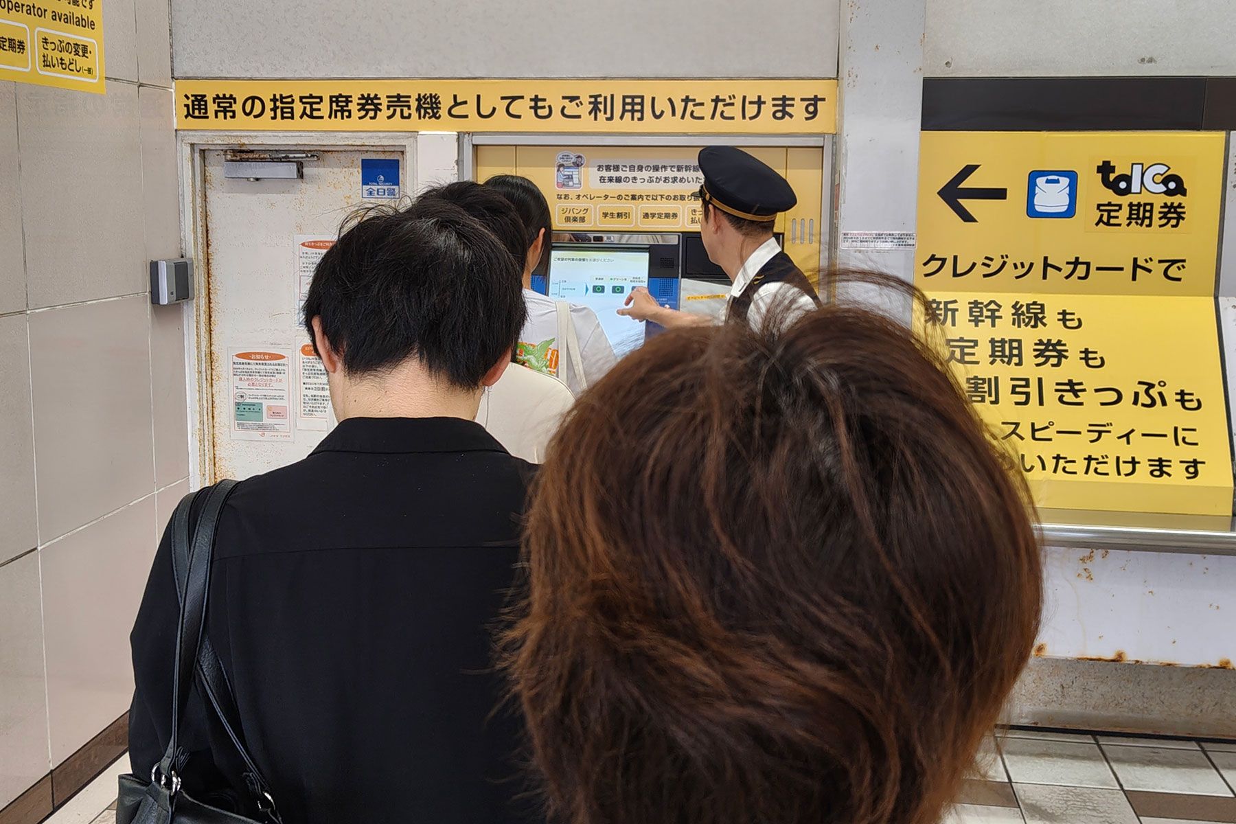 ピーク時の沼津駅では駅員が指定席券売機の操作を代行して待ち時間の短縮を図る 沼津駅の指定席券売機の様子。5〜6人ほどの行列ができており、先頭では駅員が購入希望者から行き先を聞いて操作を代行している。